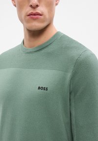 Zelený pletený sveter s rebrovanou štruktúrou, okrúhlym výstrihom a čiernym logom "BOSS" na hrudi. Materiál vyzerá mäkko a pružne.