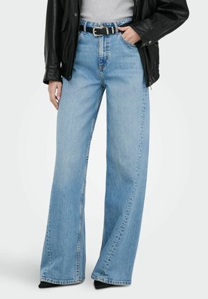 Žena má na sebe svetlomodré džínsy s širokými nohavicami, čierny opasok, sivý top a čiernu koženú bundu, jednu ruku má v vrecku.