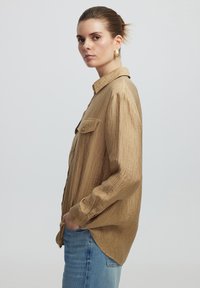 Beige Hemd aus strukturiertem Stoff mit Kragen, Brusttaschen und langen Ärmeln, kombiniert mit hellblauen Jeans.