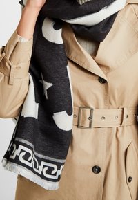 Personne portant un trench-coat beige ceinturé et une écharpe noire et blanche à motifs étoiles et bordures géométriques.