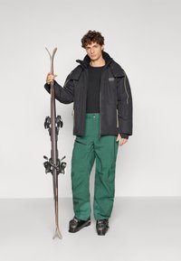 Juoda slidinėjimo striukė su geltonais akcentais, derinama su žaliais slidinėjimo kelnėmis. Modelis laiko medines slides su juodomis sagtimis, dėvi juodus slidinėjimo batus.