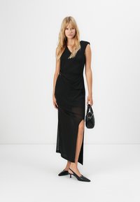 Kvinne iført en ermeløs svart kjole med sidesplit, holder en svart håndveske og har på seg svarte slingback hæler, stående mot en hvit bakgrunn.