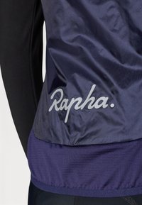 Mornarska biciklistička jakna s glatkom, sjajnom vanjštinom, elastičnim donjim rubom i sivim logom "Rapha." blizu dna, kombinirana s crnom majicom dugih rukava.