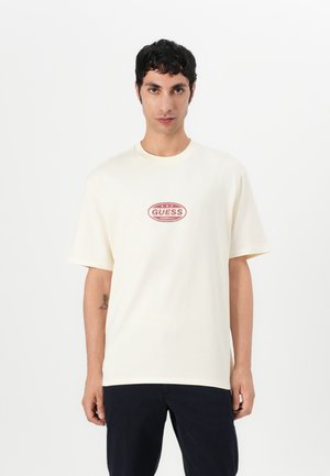 Cream cotton t-shirt with a round neck and short sleeves. Features a red oval logo with the word "GUESS" and "ORIGINALS" beneath it.