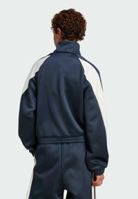 Navy blue tracksuit jacket with a high collar, white side panels, and ribbed cuffs. Smooth fabric with a relaxed fit and elastic waistband.