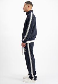 Marineblauwe trainingspak van zacht materiaal, met witte zijstrepen en logo-detail. Draagt met witte sneakers; ontworpen met een ritssluiting bij de kraag.