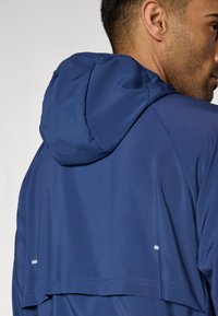 Μπλε ναυτικό μπουφάν με κουκούλα, με λεία υφή και άνετη εφαρμογή. Διαθέτει αντανακλαστικές λεπτομέρει ες κοντά στο τελείωμα για ορατότητα.