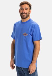 Blaues Baumwoll-T-Shirt mit rundem Ausschnitt, kurzen Ärmeln und einem rautenförmigen Grafikdesign mit einem Augensymbol auf der Brust.