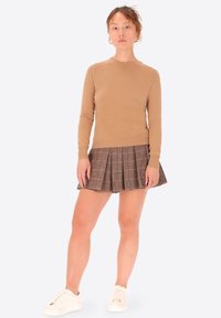 Maglione aderente marrone chiaro abbinato a una gonna plissettata a quadri nei toni del marrone scuro e beige, indossato con sneakers bianche.