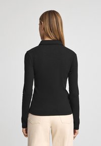 Polo a maniche lunghe nera con colletto, caratterizzata da una progettazione aderente e una texture liscia. Indossata con pantaloni beige chiari.