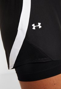 Bermudas deportivas negras con una franja lateral blanca y un panel de malla, que presentan un logo blanco de Under Armour en el muslo, llevadas en la pierna de una persona.