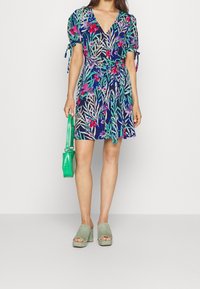 Květinové zavinovací šaty s nabíranými rukávy v námořnické modré, zelené a růžové barvě. Nošeny se zelenou kabelkou a zelenými mulemi s blokovým podpatkem.