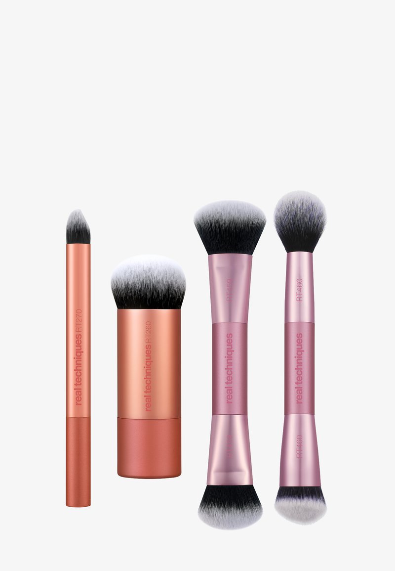 Quatre pinceaux de maquillage à double extrémité et à extrémité simple, avec des poils noirs et blancs et des manches en or rose et rose, étiquetés "real techniques".