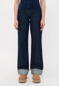 Person som har på seg mørkeblå vidde jeans med oppbrettede nederkant, brune platåsko, og en marineblå knappeløs topp, stående mot en hvit bakgrunn.