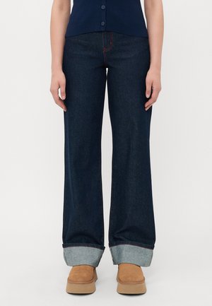 Person som har på seg mørkeblå vidde jeans med oppbrettede nederkant, brune platåsko, og en marineblå knappeløs topp, stående mot en hvit bakgrunn.