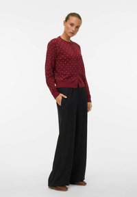 Femme avec les cheveux attachés porte un cardigan bordeaux à pois, un pantalon noir large et des chaussures marron, se tenant avec une main dans la poche.