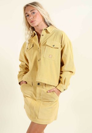 Femme portant une veste en velours côtelé jaune avec poches avant et une jupe assortie, les mains dans les poches, sur un fond clair uni.