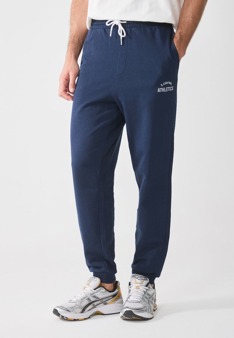 Marineblå idrettsjoggesko med elastisk liv, hvit snor, sidelommer og brodert "S. CENTRAL ATHLETICS"-detalj.