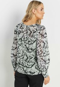 Blusa verde chiaro con un motivo astratto in bianco e nero, maniche lunghe a sbuffo trasparenti e chiusura a chiave sul retro, realizzata in un tessuto leggero.