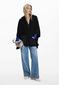 Fekete kapucnis poncsó elöl cipzárral, nagy zsebekkel és húzózsinórral. Széles szárú kék farmerral és mintás táskával párosítva.