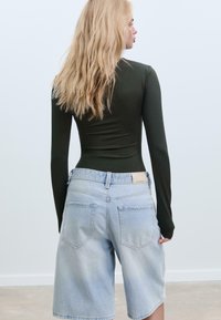 Langärmliges, dunkelgrünes, enganliegendes Oberteil kombiniert mit hellblauen Jeansshorts, die über Front- und Gesäßtaschen, lockere Passform und ausgefranste Säume verfügen.