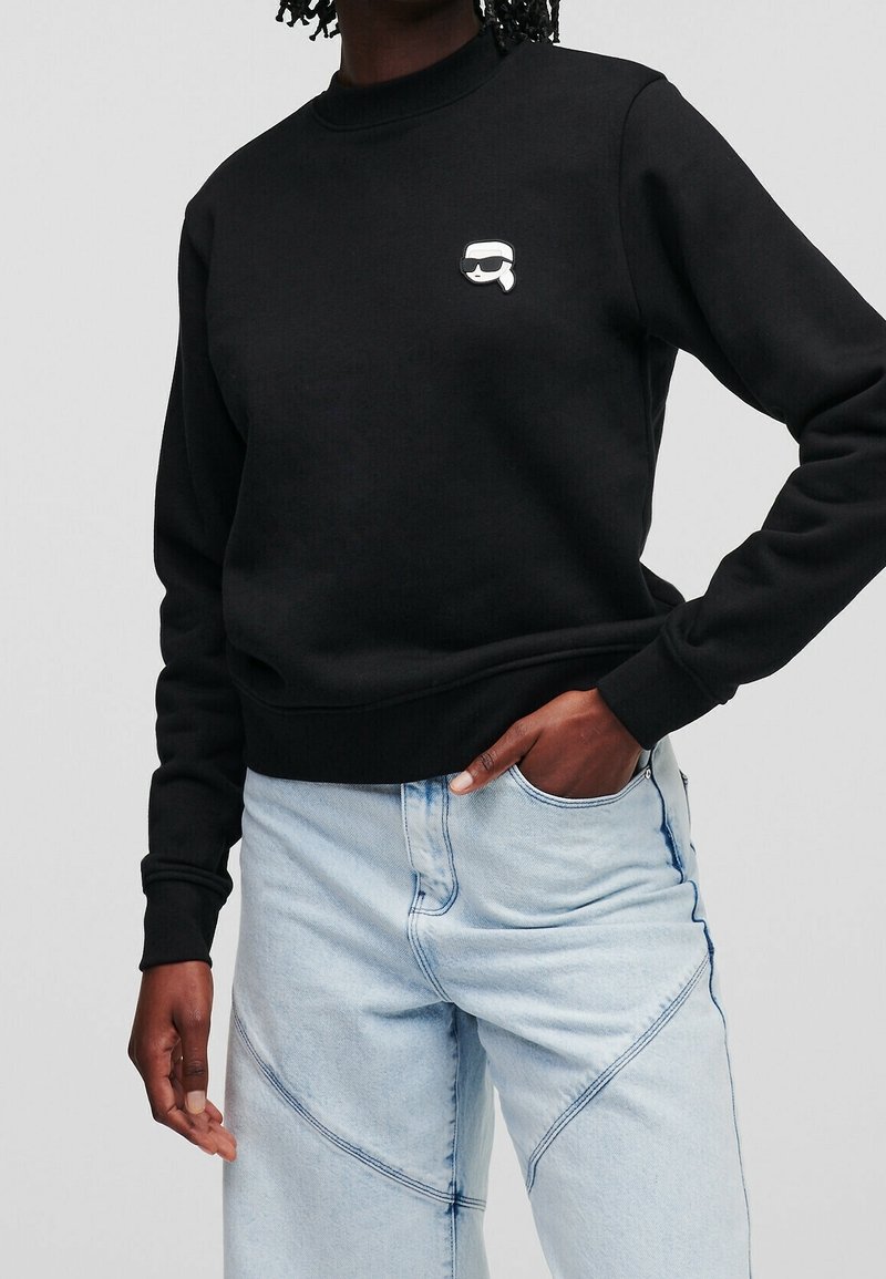 Czarny sweter z okrągłym dekoltem, ściągaczami na rękawach i małym białym nadrukiem na klatce piersiowej. Zestawiony z jasnoniebieskimi dżinsami.