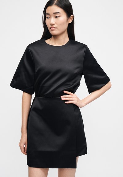 Kvinne iført en kort svart satengkjole med vide ermer, stående med en hånd på hoften mot en ensfarget hvit bakgrunn.