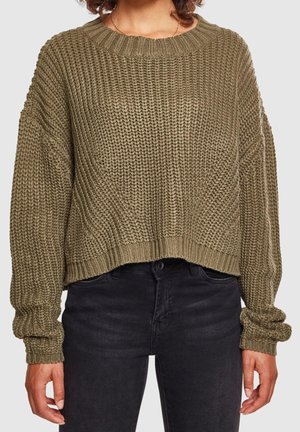 Personne portant un pull en grosse maille vert olive à manches longues et un jean noir taille haute, debout devant un fond clair uni.
