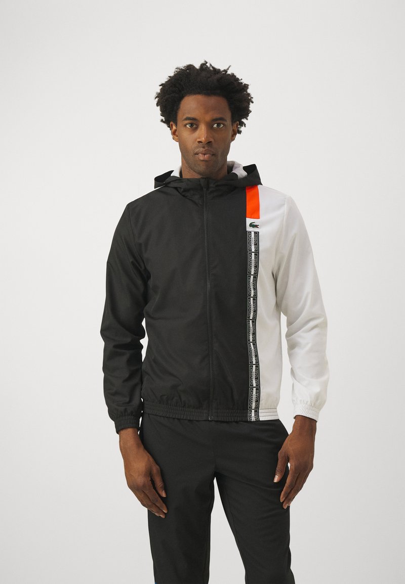 Uomo che indossa una giacca a zip con cappuccio bianca e nera, con una striscia arancione e nastro verticale con un motivo sul lato destro, abbinata a pantaloni neri.