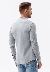 Camisa de manga larga gris con puños abotonados, presenta un dobladillo redondeado y un collar. La tela tiene una textura suave y lisa. Combinada con jeans azules.