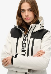 Weiße und schwarze wasserdichte Jacke mit Reißverschluss, verstellbarer Kapuze, auffälligem Logo und Reißverschlusstaschen. Weich texturierte Bündchen sind sichtbar.