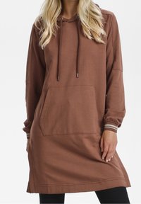 Abito cappuccio marrone realizzato in tessuto morbido, con una tasca frontale, maniche lunghe e dettagli a righe sui polsini. Cappuccio regolabile con lacci.