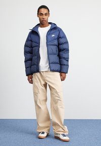 Jovem vestido com uma jaqueta acolchoada Nike azul-marinho, camiseta branca, calças de carga bege e ténis brancos com atacadores azul-marinho, em pé sobre um tapete azul.