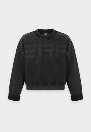 Homme portant un sweat-shirt noir et un pantalon cargo camouflage, debout devant un fond clair uni.