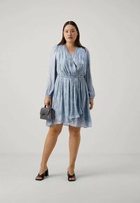 Femme portant une robe portefeuille bleu clair à manches longues, tenant un petit sac à main gris et portant des sandales à talons blocs noires ouvertes au bout, se tenant contre un fond uni.