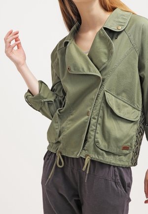 Femme portant une veste décontractée verte avec de grandes poches avant et des cordons, associée à un pantalon ample et sombre, posant avec une main levée.