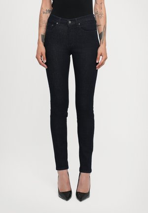 Skinny denim jeans in dark blue with a smooth texture, featuring front pockets and a five-button fly. Worn with pointed black heels.
