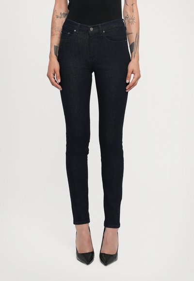 Jeans de denim ajustados en azul oscuro con una textura suave, que cuentan con bolsillos frontales y una bragueta de cinco botones. Llevados con tacones negros puntiagudos.