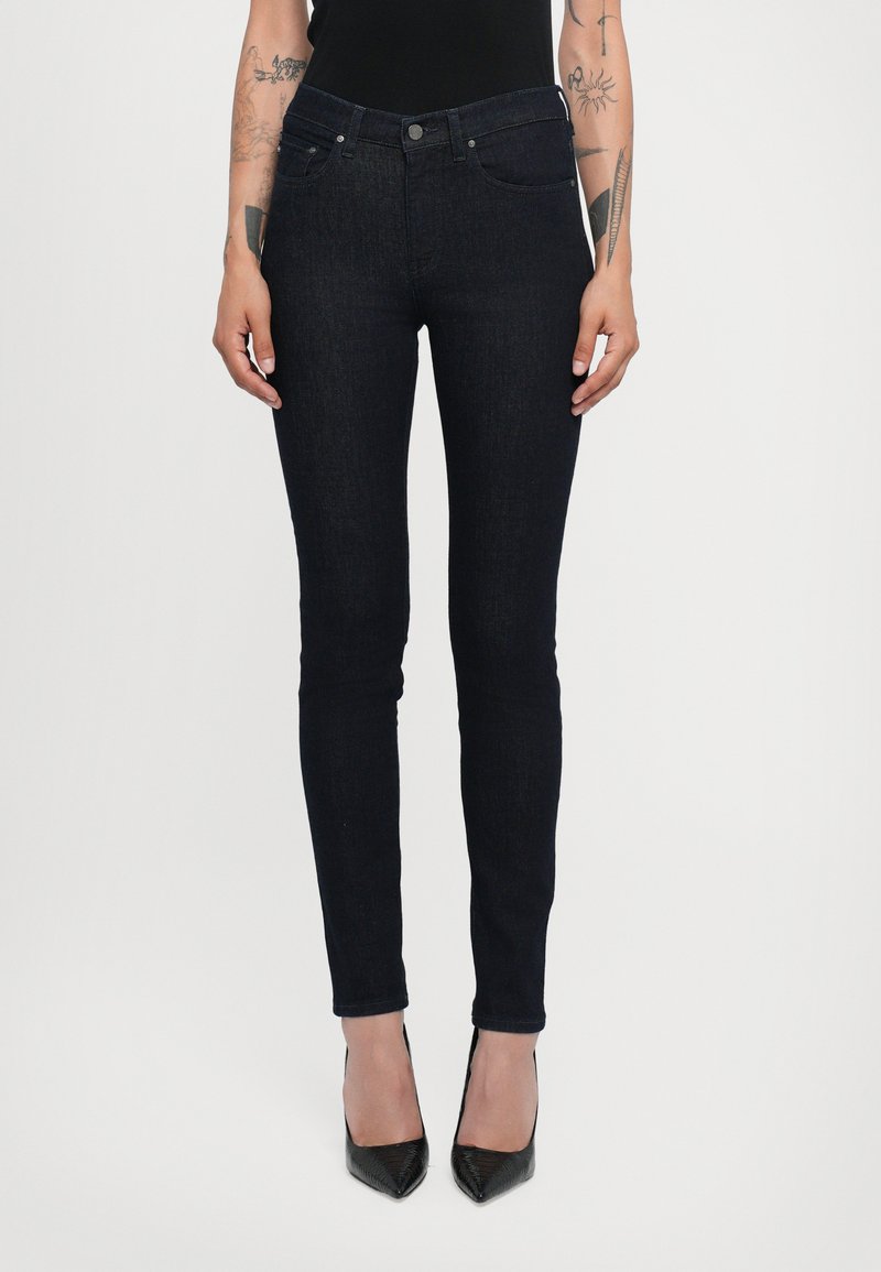Dünne Jeans aus Denim in dunkelblau mit glatter Oberfläche, ausgestattet mit Fronttaschen und einem Fünf-Knopf-Verschluss. Tragen mit spitzen schwarzen High Heels.