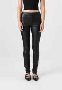 Leggings neri in ecopelle con vestibilità slim, caratterizzati da un alto punto vita e una texture liscia e lucida, abbinati a scarpe nere con punta affusolata.