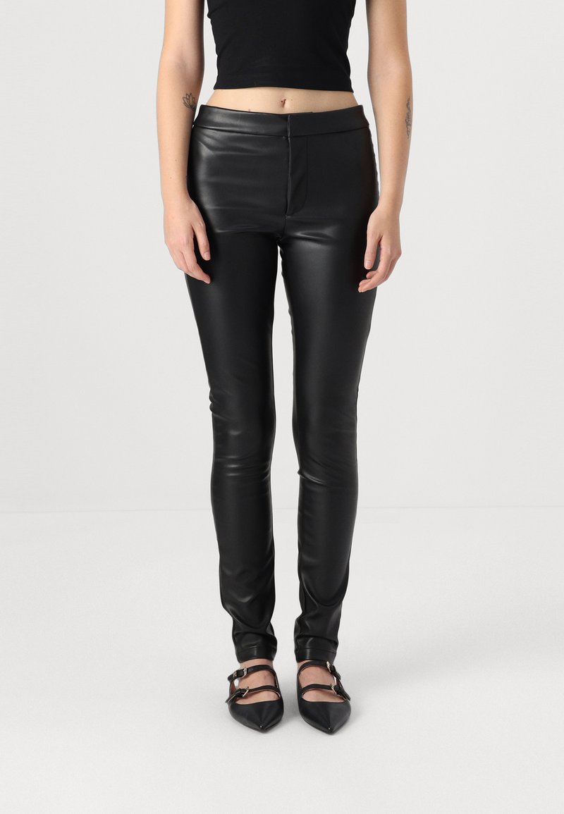 Leggings neri in ecopelle con vestibilità slim, caratterizzati da un alto punto vita e una texture liscia e lucida, abbinati a scarpe nere con punta affusolata.