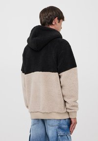 Giacca in pile con cappuccio caratterizzata da un design color block, con una sezione superiore nera e una inferiore beige, realizzata in un tessuto morbido e texturizzato.