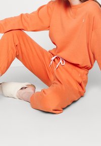 Osoba sedící v jasně oranžové mikině a odpovídajících teplácích s bílou šňůrkou a krémově zbarvenými huňatými pantoflemi.