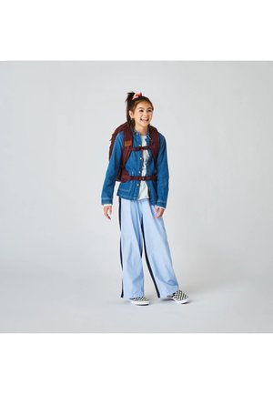 Lächelndes Mädchen mit einer Jeansjacke, hellblauen gestreiften Hosen, karierten Slip-on-Schuhen und einem dunkelroten Rucksack vor einem schlichten grauen Hintergrund.