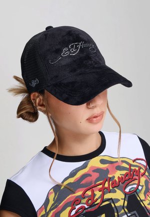 Kobieta nosząca czarną czapkę Ed Hardy z logo z kryształków oraz białą koszulkę Ed Hardy z kolorowym wzorem tygrysa i czerwonym napisem.