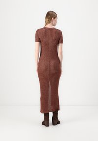 Rippendes, rostfarbiges Strickkleid mit kurzen Ärmeln, taillierter Form und einem seitlichen Schlitz. Kombiniert mit braunen Stiefeln mit Absatz.