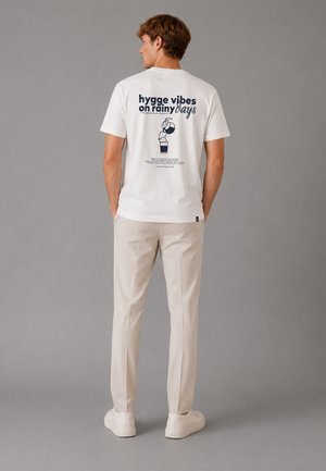 Homme portant un t-shirt blanc avec le texte "hygge vibes on rainy days" et un visuel de café versé dans le dos, pantalon beige et baskets blanches.