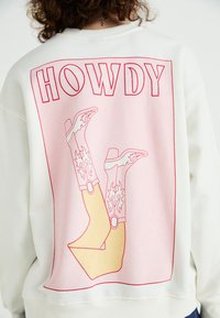 Witte sweater met een grote roze graphic met de tekst "HOWDY" en geïllustreerde cowboylaarzen met decoratieve vlammen, omrand door rood.