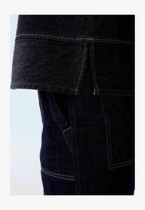 Tmavě modrý denimový kousek s volným lemem, kontrastním žlutým prošíváním a viditelnou kapsou se strukturovaným tvarem.