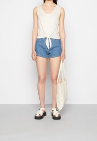 Světle béžový bezrukávový top s mašlí v pase, kombinovaný s džínovými šortkami s ohrnutým lemem a bílými sandály na nártu. Béžová taška.